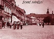 Na snímku je znázorněn stav náměstí na počátku 20. století v době dopoledního prodeje v obchodech, které jsou rozesety po obou stranách náměstí